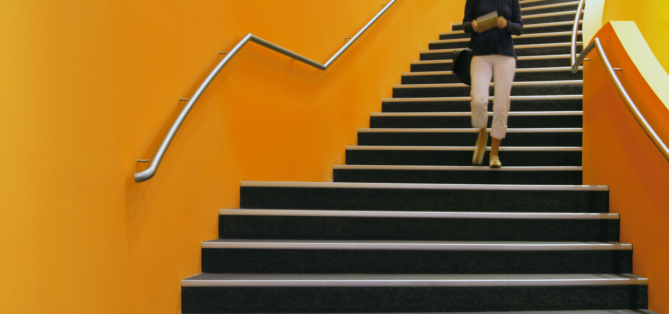 A young woman carrying a book in both hands descends a curved staircase in a public building.