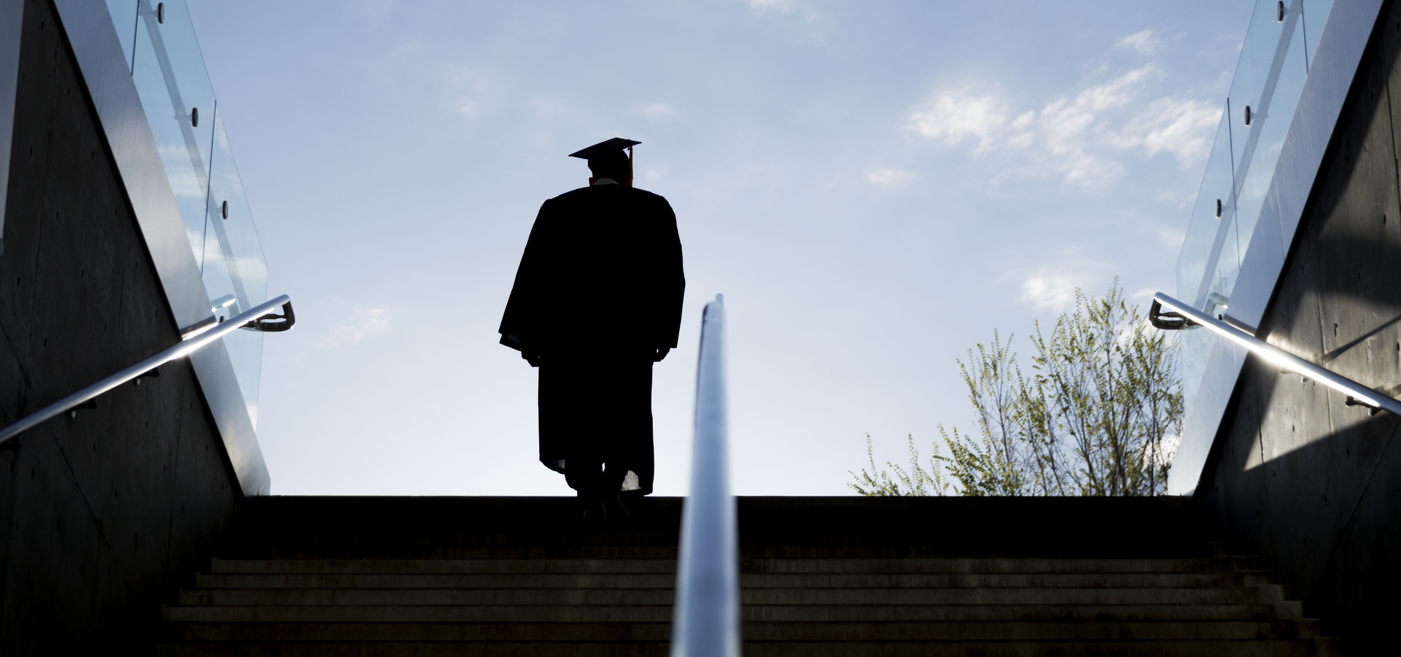 A young graduate in cap and gown cuts a silhouette in the morning sky as they ascend a staircase. 